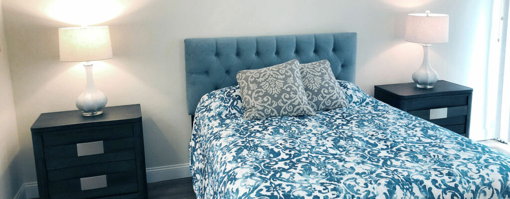 Large bed with a blue paisley comforter and throw pillows, blue suede headboard and two side tables with lamps.